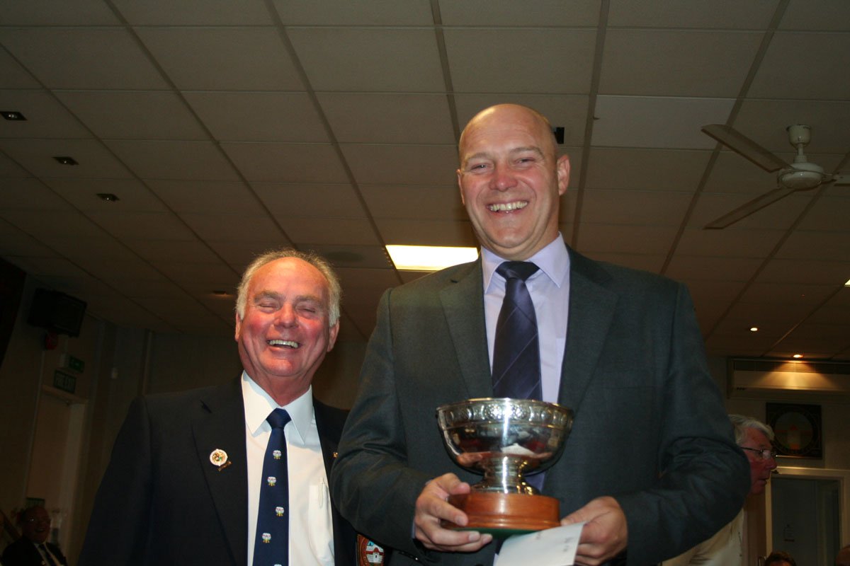 Two men in suits smiling at an indoor event, with the man on the right holding a silver trophy and a piece of paper.