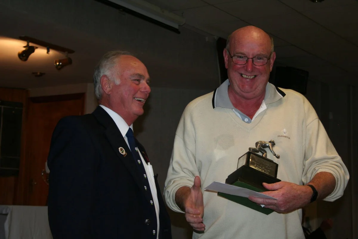 Two smiling men at an award ceremony; one holding a trophy of a dog, the other in a suit.