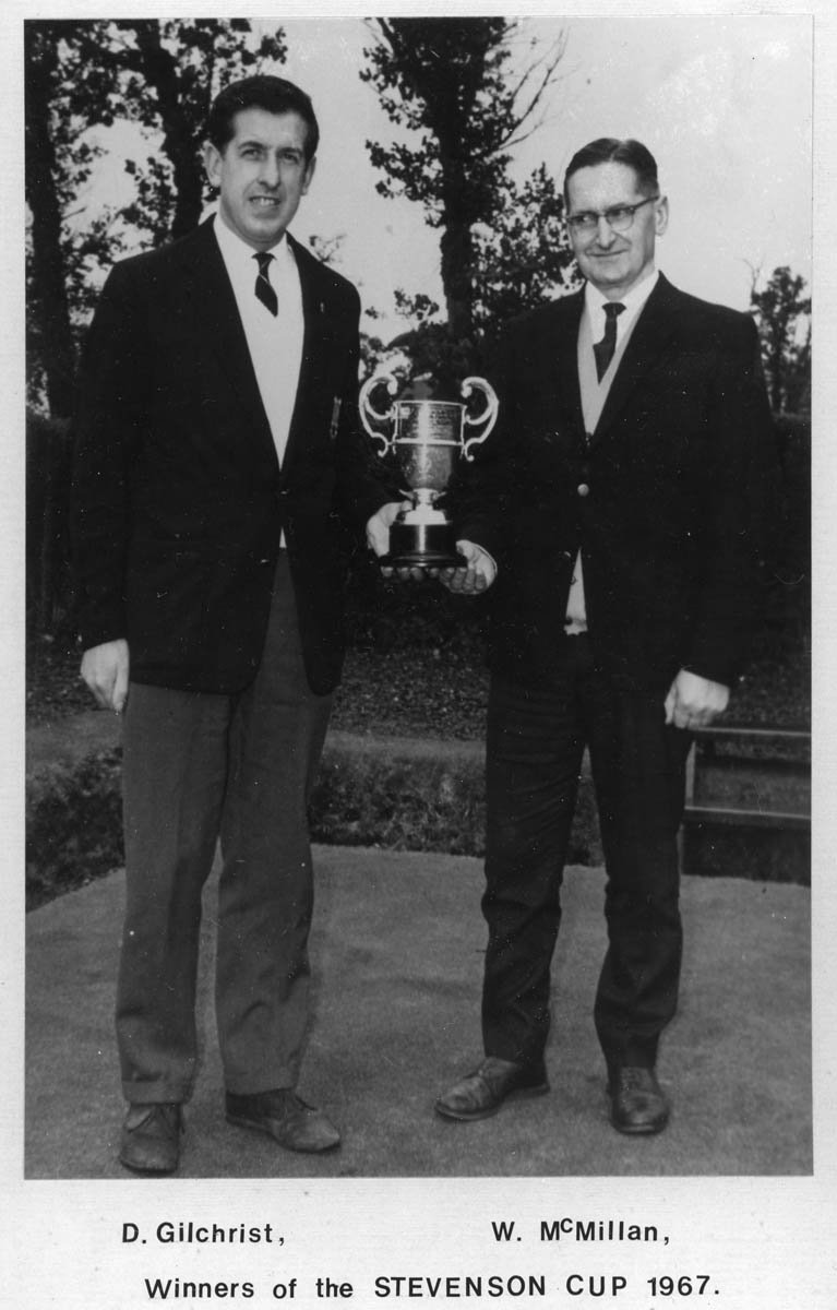 Two men in suits standing outdoors, holding a large trophy cup, with trees in the background, celebrating their win of the Stevenson Cup in 1967.