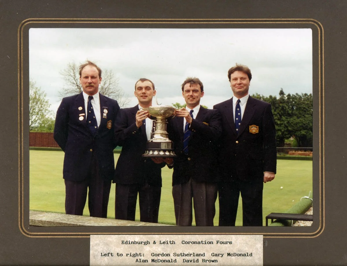 Four men dressed in formal suits, standing outdoors on a golf course, holding a large trophy cup together. Two of the men are wearing badges. The background shows trees and a cloudy sky.