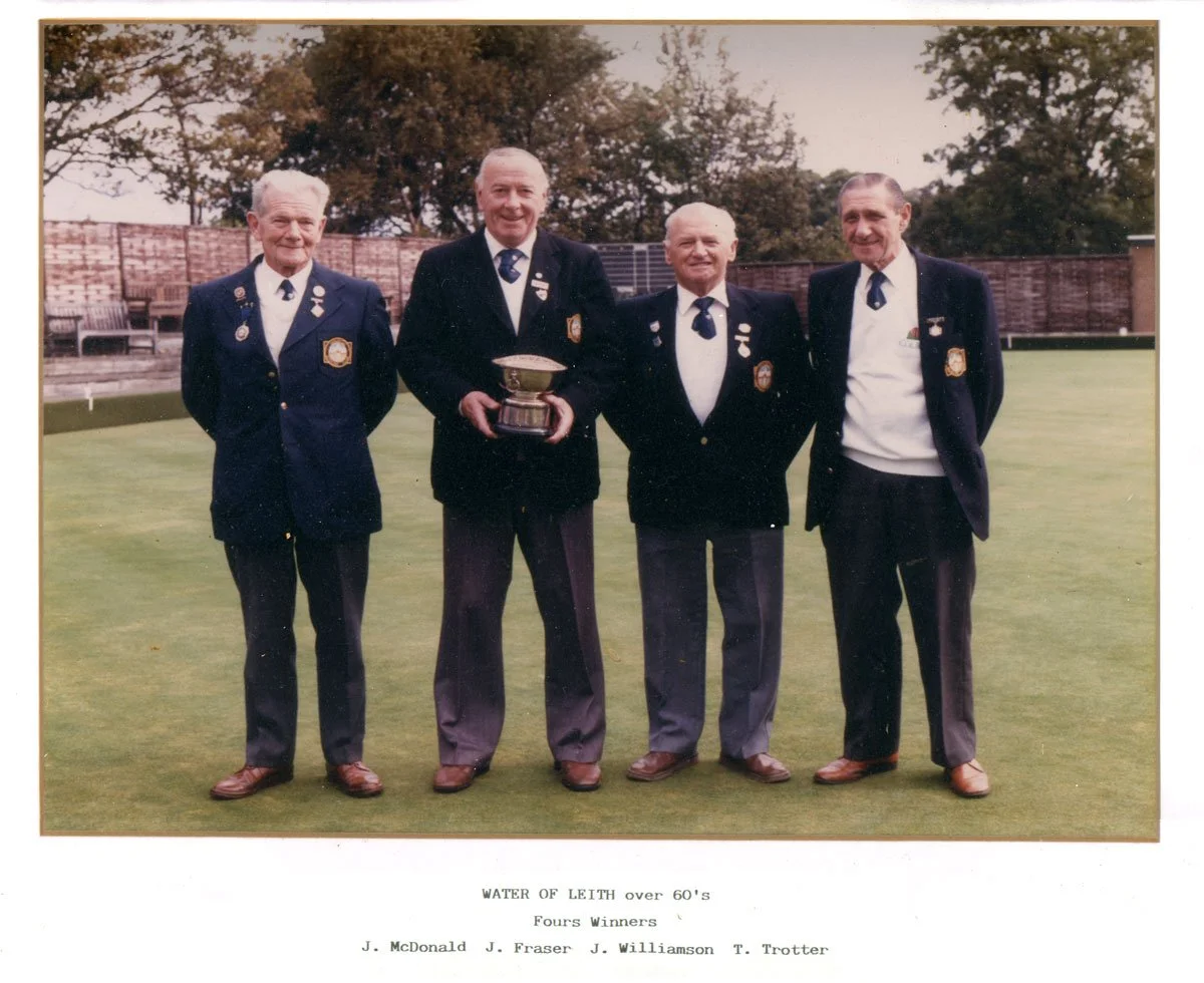 Four elderly men wearing blazers with medals standing on a grass field, one holding a trophy, in an outdoor setting with trees and a wooden fence in the background.