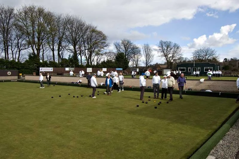 People playing lawn bowls on a green field under a cloudy sky, with trees and a building in the background.