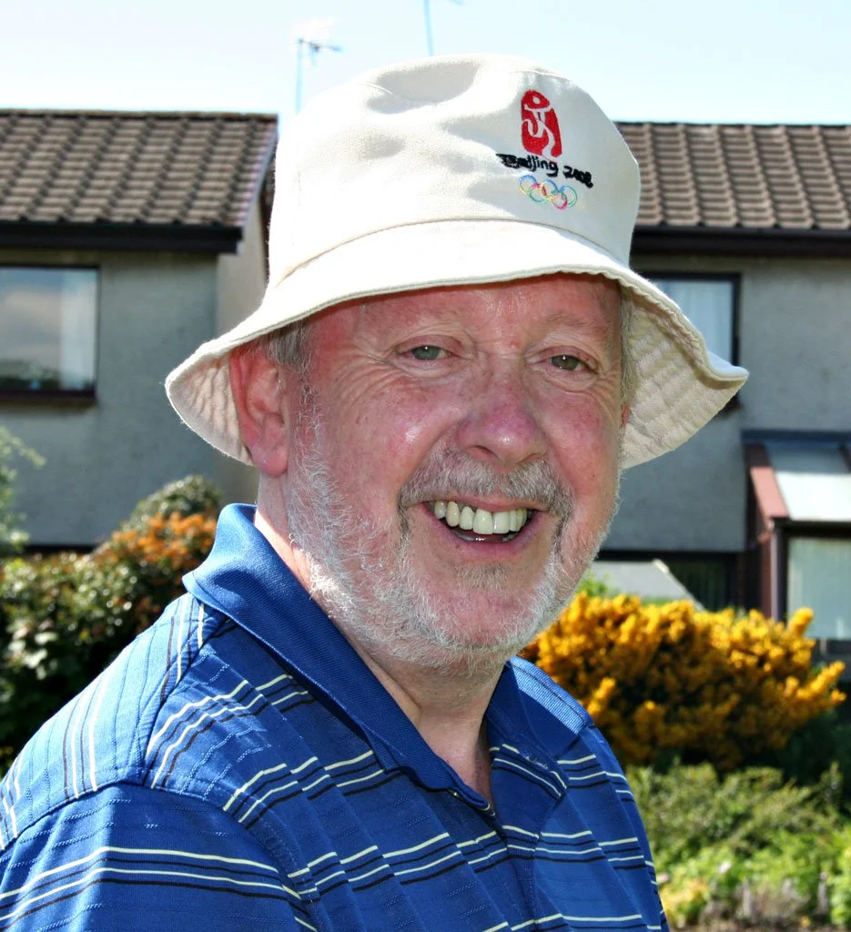 Smiling man outdoors wearing a wide-brimmed hat with the Beijing 2022 Winter Olympics logo, blue striped shirt, and standing in front of a house with flowers and shrubs.