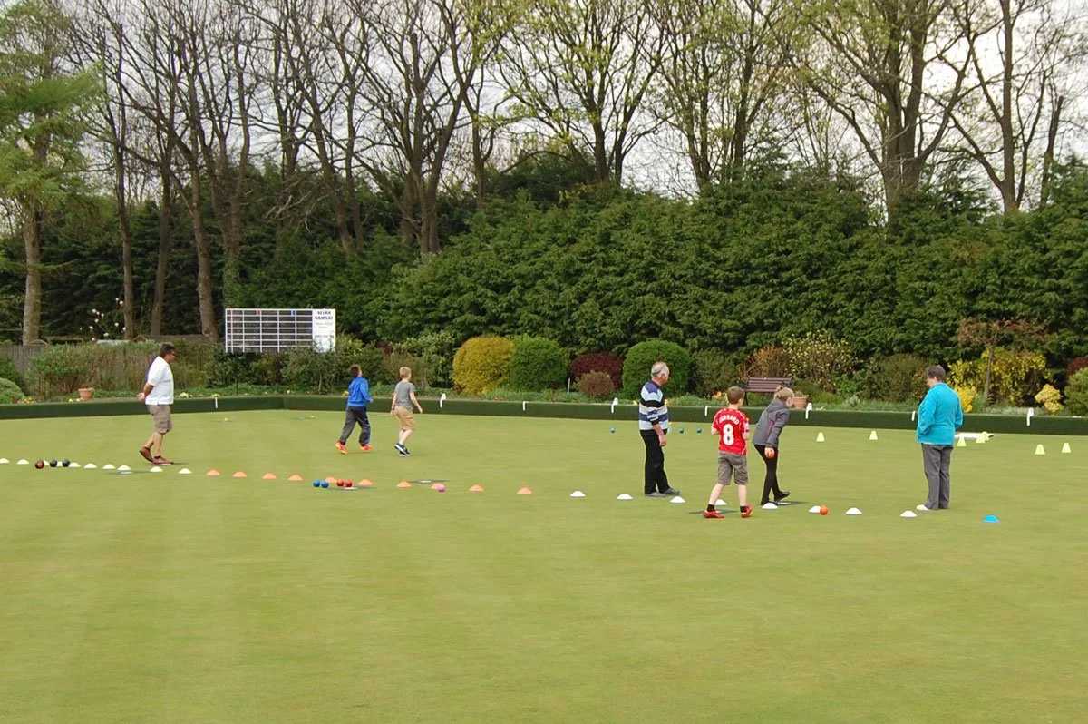 People practicing lawn bowls on a green field with trees and bushes in the background.