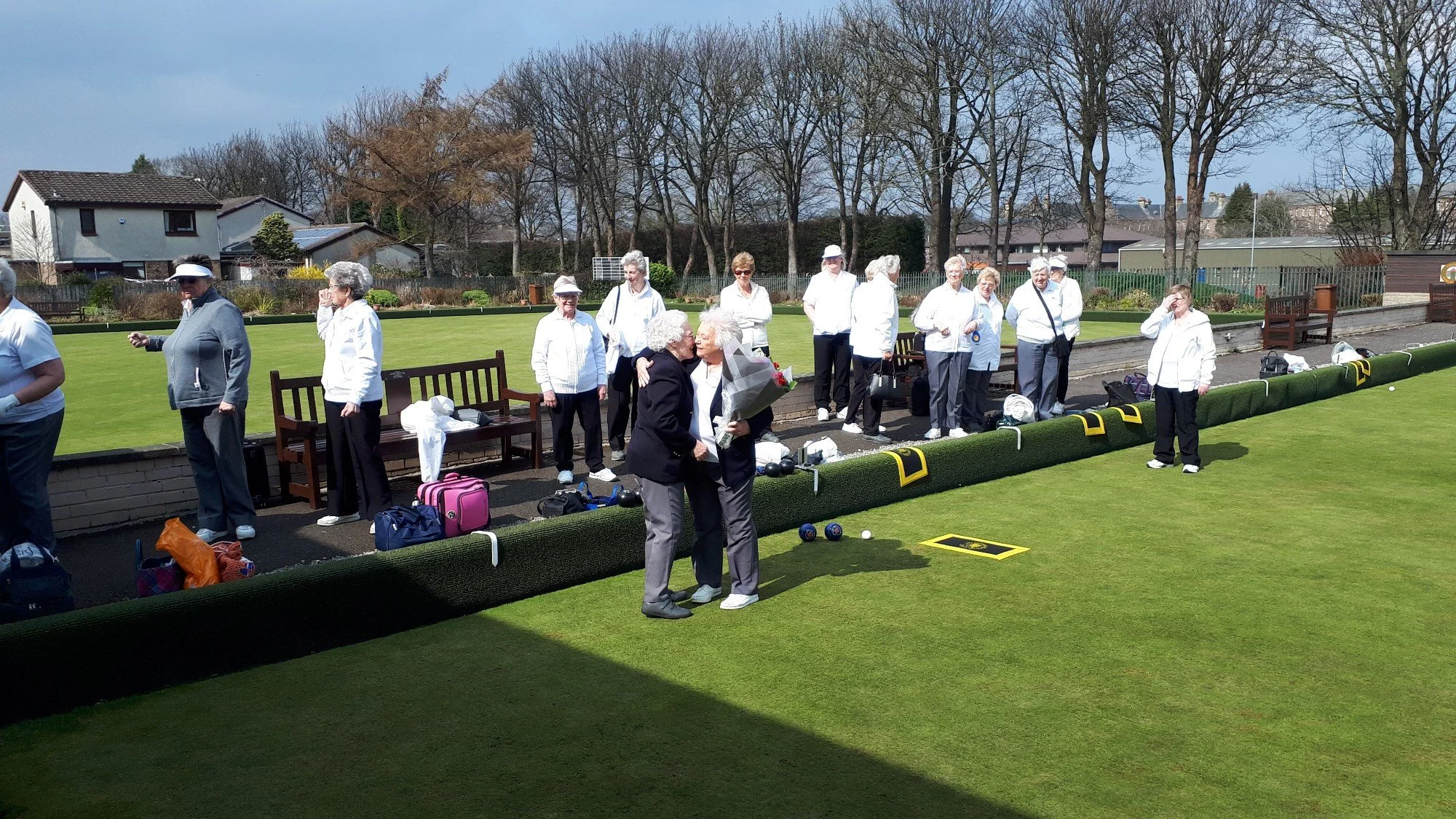 A group of elderly women dressed in white and black, some with hats or sunglasses, gathered on a lawn bowling green with equipment and bags around. Two women in the foreground are embracing and smiling, holding flowers. The background shows some hous