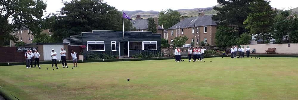 People playing lawn bowls on a green field in front of a modern building with purple flag, surrounded by houses and trees, in a hilly area.