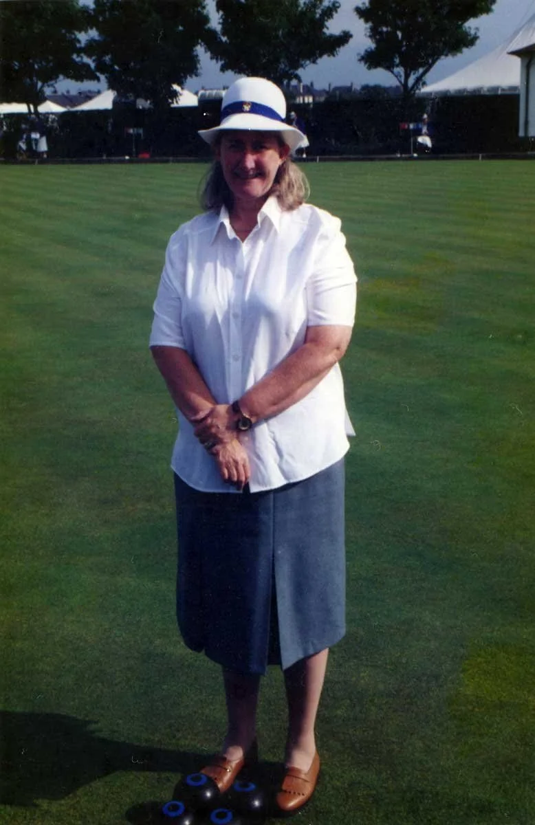 A woman standing on a golf course wearing a white shirt, a gray skirt, a white hat with a blue band, and brown shoes, with a black watch on her left wrist, and a few golf balls with blue rings at her feet.