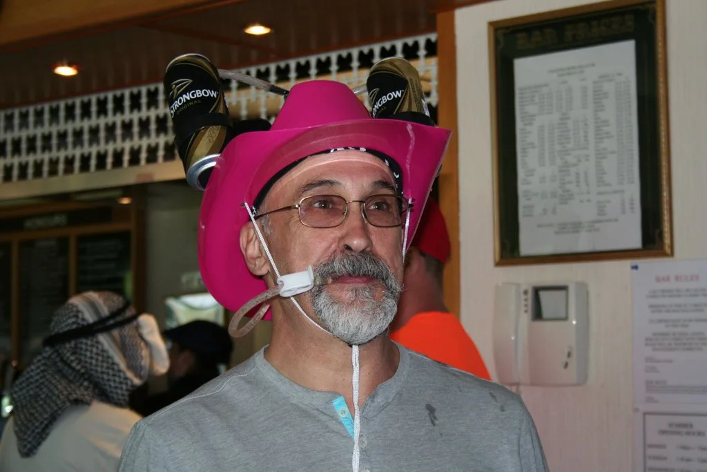 A man with a gray beard and glasses wearing a pink cowboy hat adorned with a plastic bottle holder, two Strongbow cider bottles, and a straw. He is indoors at a casual gathering.