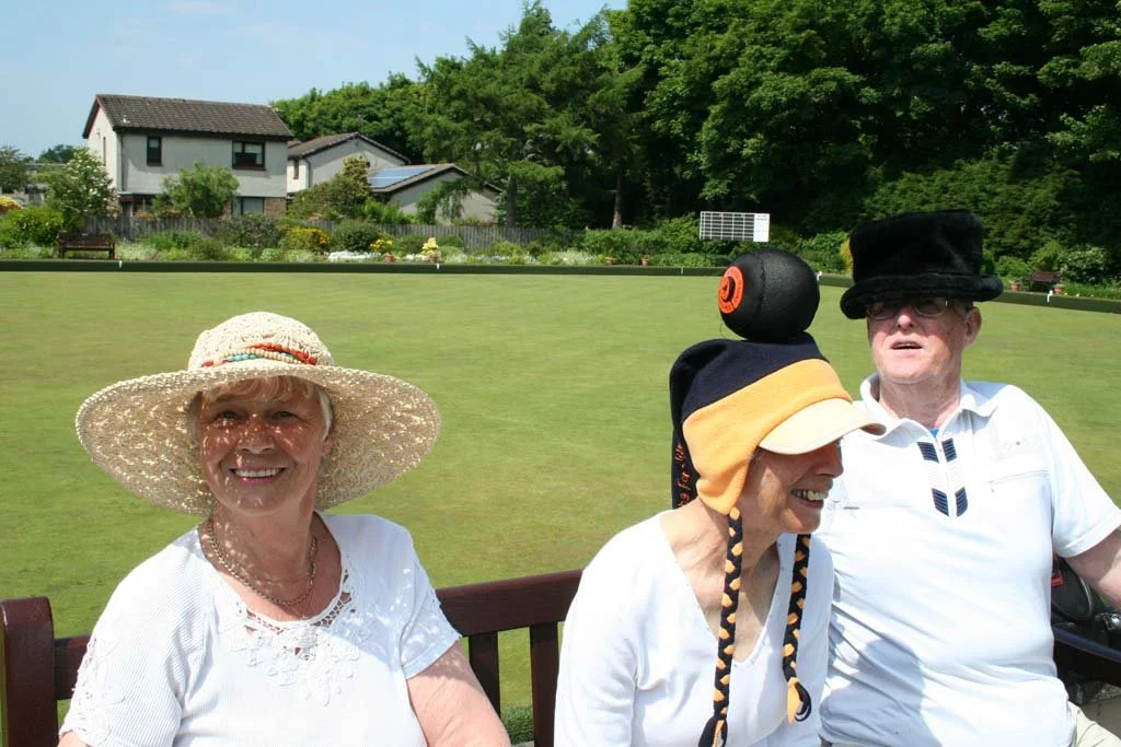 Three elderly women sitting on a bench outdoors on a sunny day, wearing hats, with a grassy area and houses in the background.