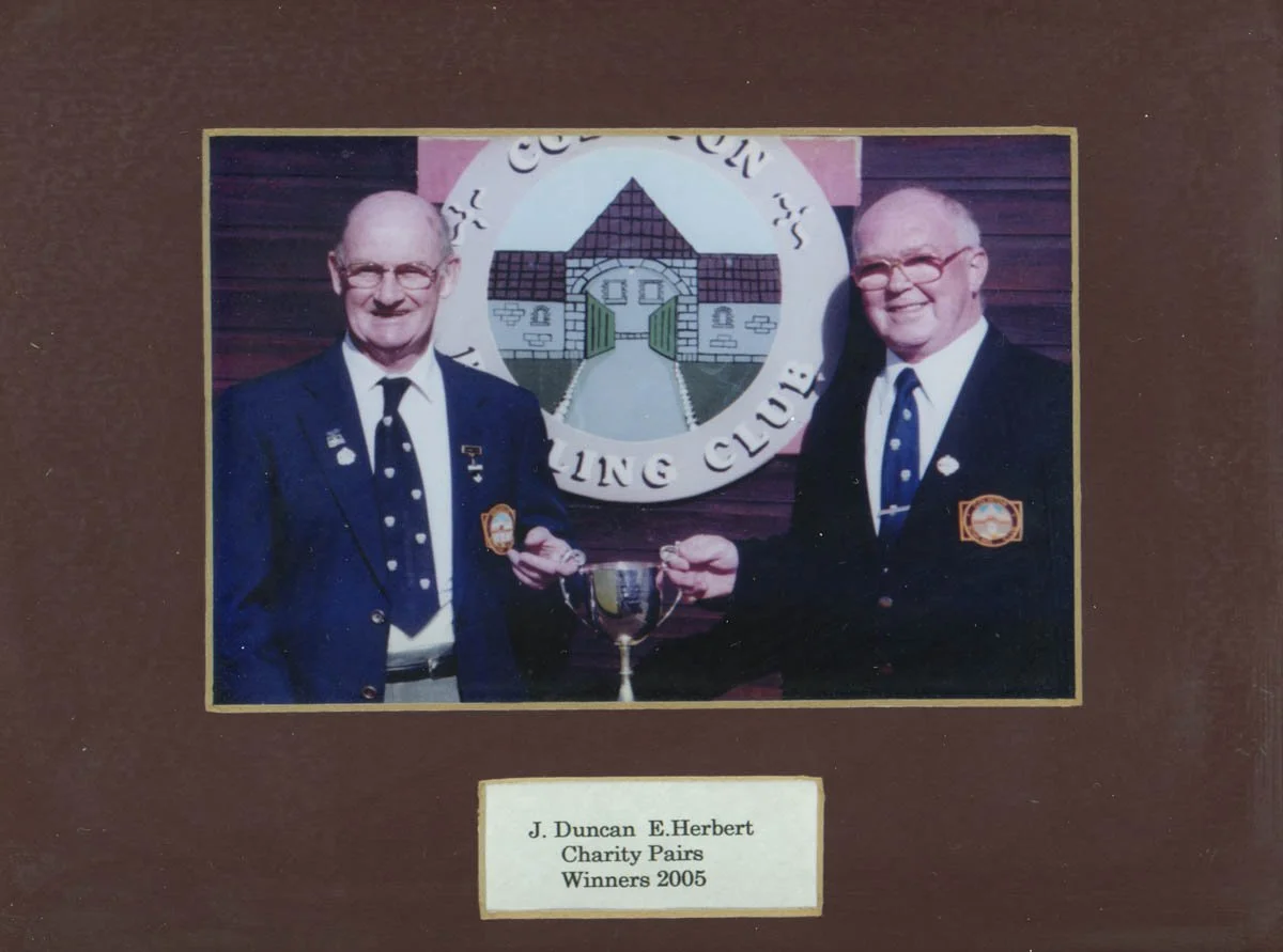 Two men in suits shaking hands and holding a trophy in front of a sign that reads "Golf Club" with a logo of a building with a gate. A plaque below the image reads "J. Duncan E. Herbert Charity Pairs Winners 2005."
