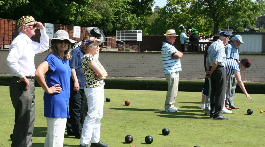 People playing lawn bowls outdoors on a sunny day with a wooden fence and trees in the background.