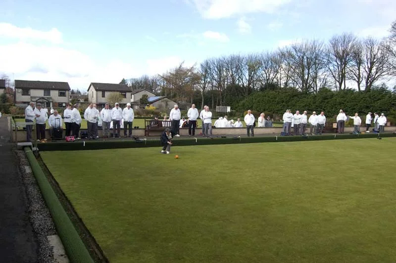 People dressed in white jackets playing or watching a game of lawn bowls on a grassy outdoor court, with houses and trees in the background.