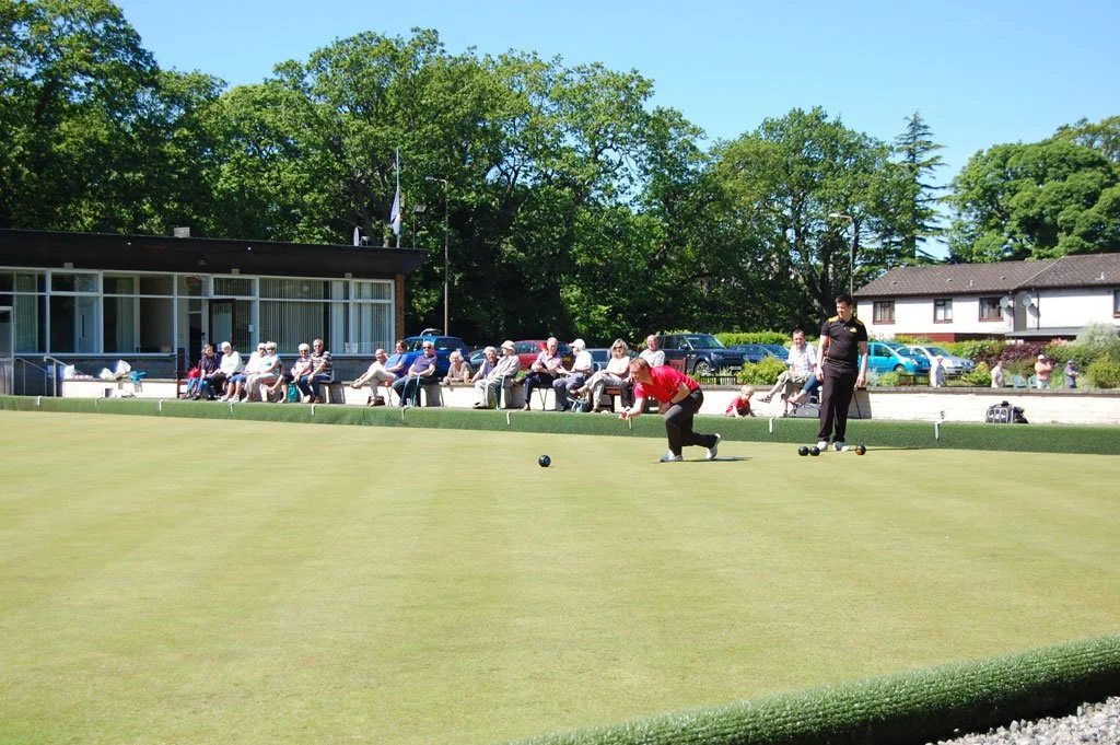 People playing lawn bowls on a green with an audience seated behind.