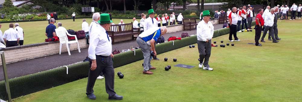 People playing lawn bowls on a green, with some waiting and observing. Many participants are wearing white shirts and green hats, typical of lawn bowls attire. A brick wall and garden area are visible in the background.