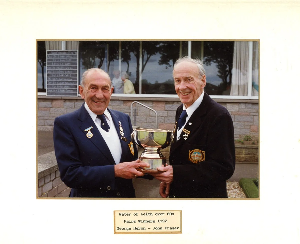 Two elderly men in suits holding a trophy in front of a building with a window and trees reflected in the glass. The man on the left is wearing a dark suit with pins and medals, and the man on the right is also in a dark suit with a pin and a badge. 