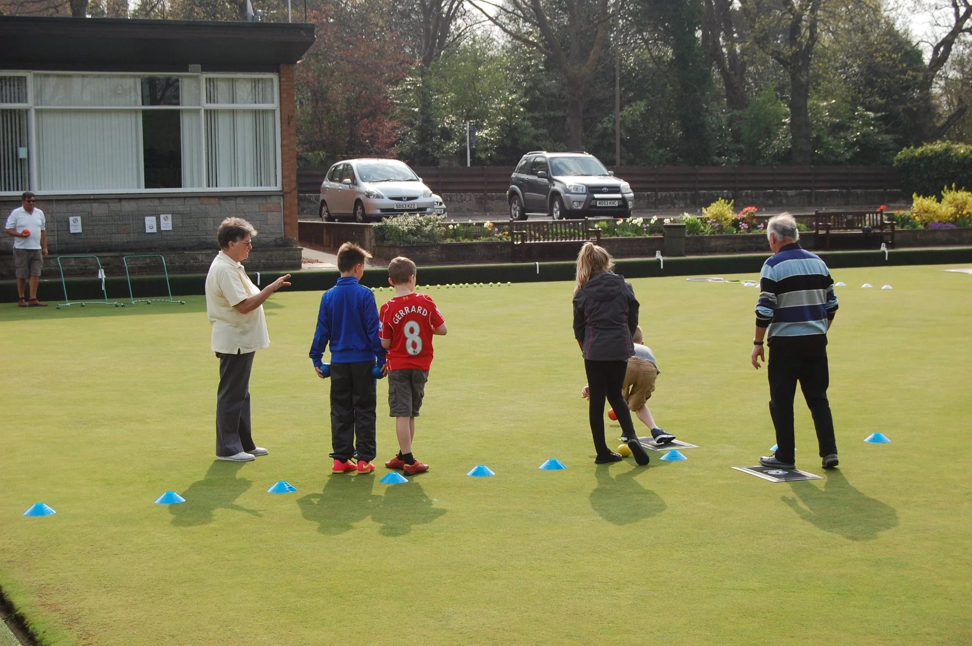 People playing lawn bowls on a green with cones, a couple of children, and adults watching or instructing. In the background, cars are parked along a street, and trees and a building are visible.
