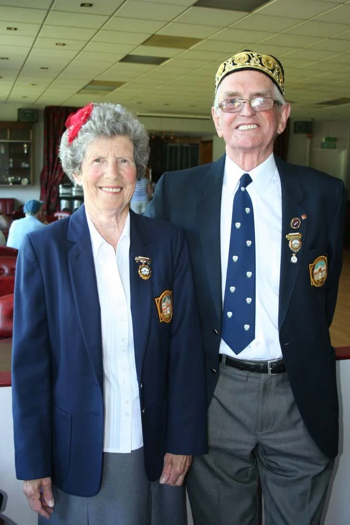 An elderly woman and man in formal attire posing together indoors. They are wearing navy blazers with pins and patches, suggesting they are part of a club or organization. The woman has gray hair, a red decorative bow, and is smiling. The man has gla