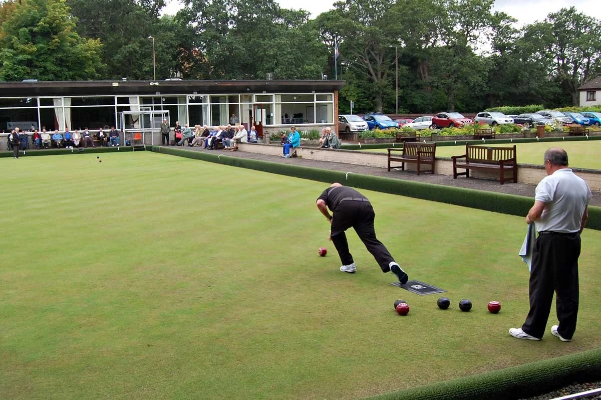 People playing lawn bowls on a green with spectators and parked cars in the background.