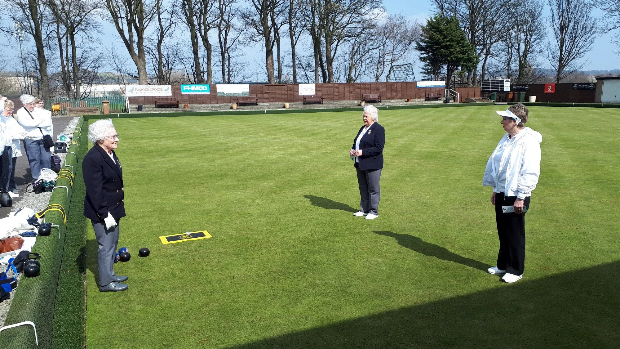 Women participating in a lawn bowling game on a sunny day in a park-like setting surrounded by trees.