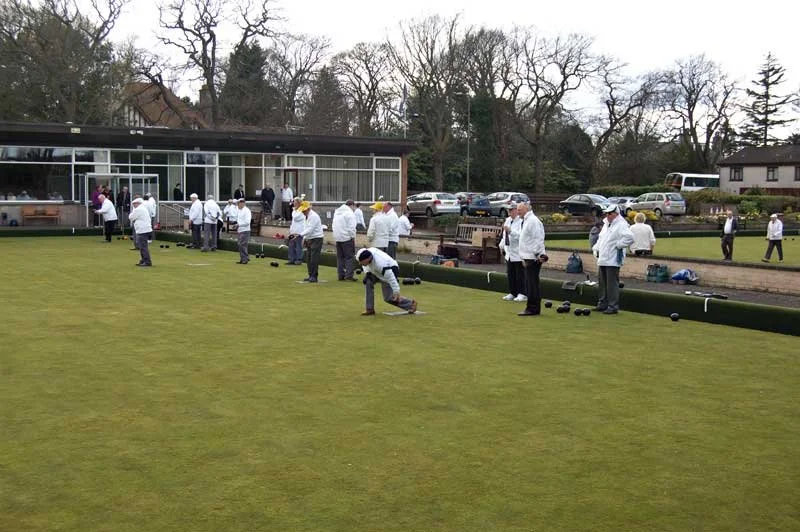 People playing lawn bowls on a green, with some preparing to bowl and others watching, on a cloudy day.