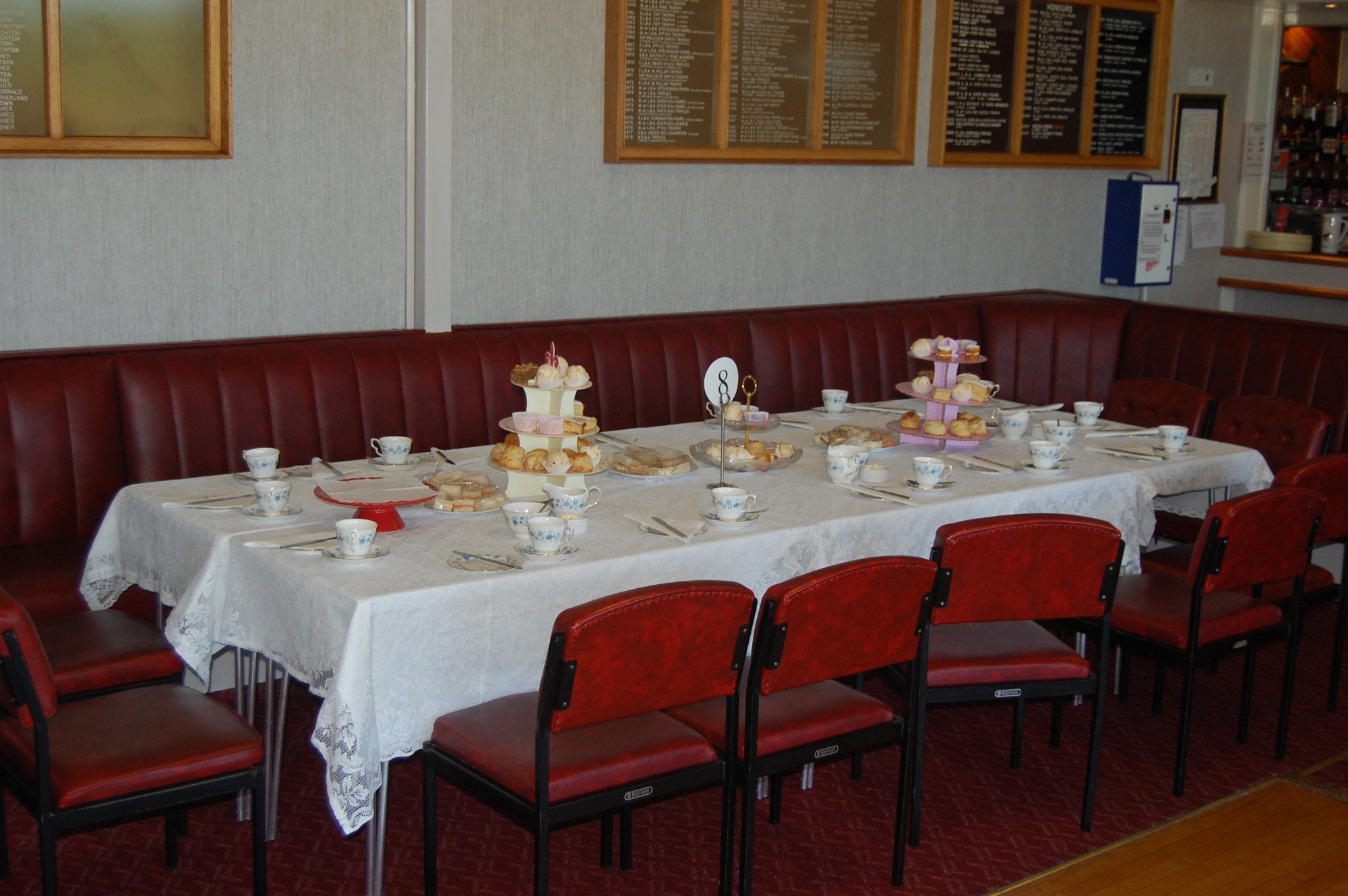 A long table set for an afternoon tea or dessert, covered with a lace tablecloth. The table has multiple cups and saucers, tiered trays filled with assorted baked goods and pastries, and a white decorative stand with cupcakes on top. Red-upholstered 