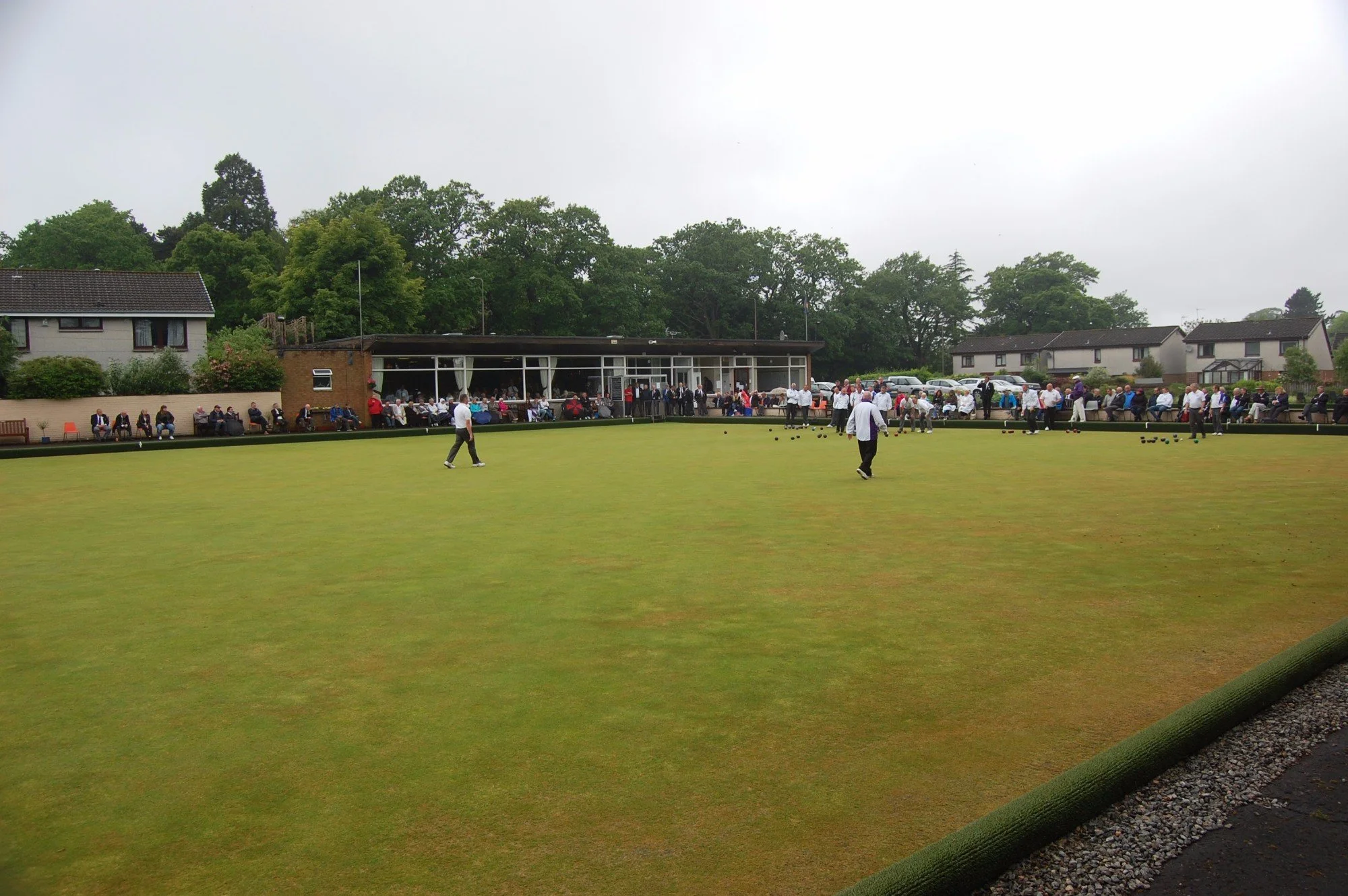 Lawn bowls game with players and spectators at the side, buildings and trees in the background on a cloudy day.