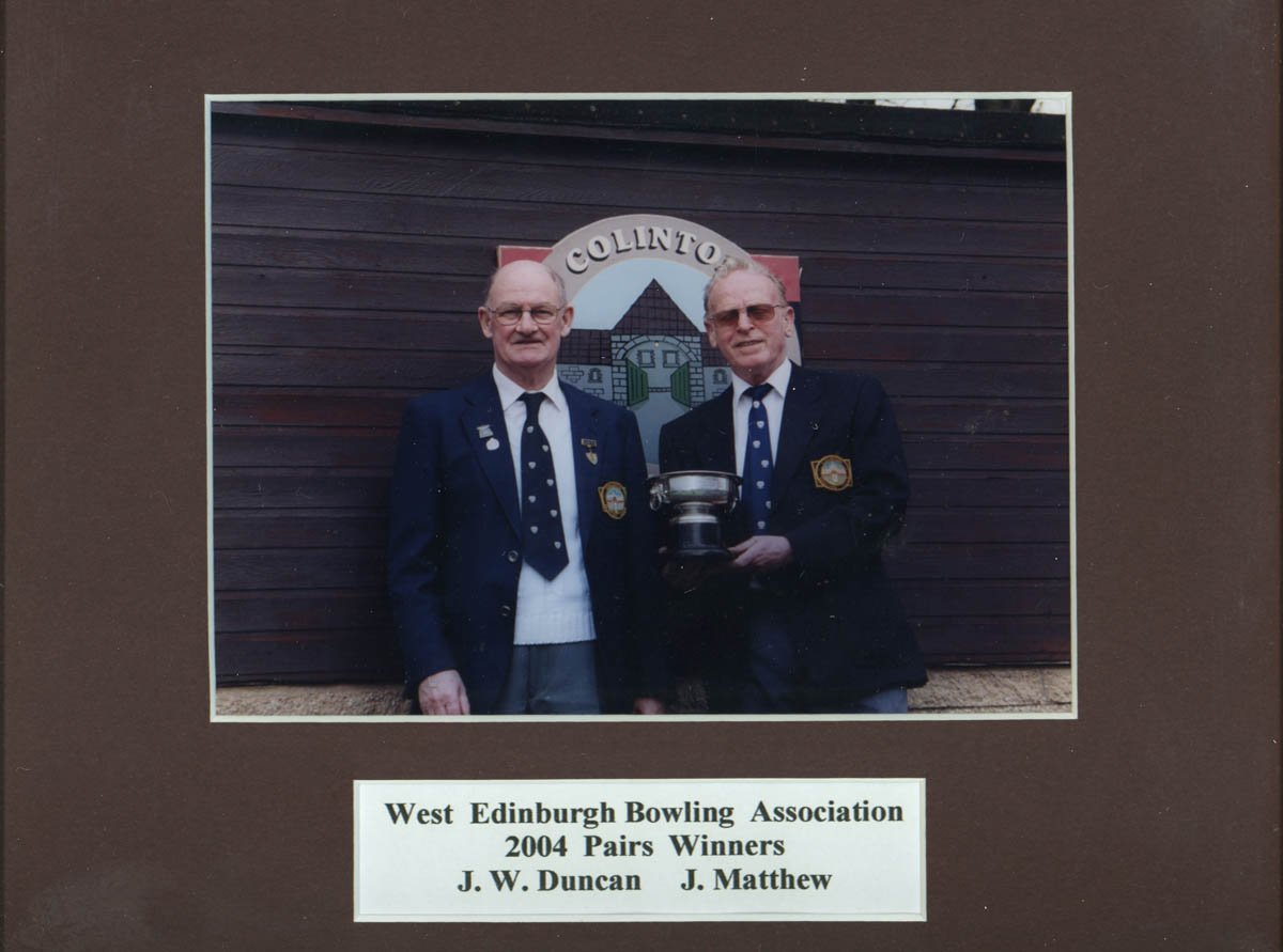 Two men in bowtie and blazer suits with badges standing in front of a wooden wall with a crest that says 'Collinton.' One man holds a trophy, and a sign below reads 'West Edinburgh Bowling Association 2004 Pairs Winners J. W. Duncan J. Matthew.'