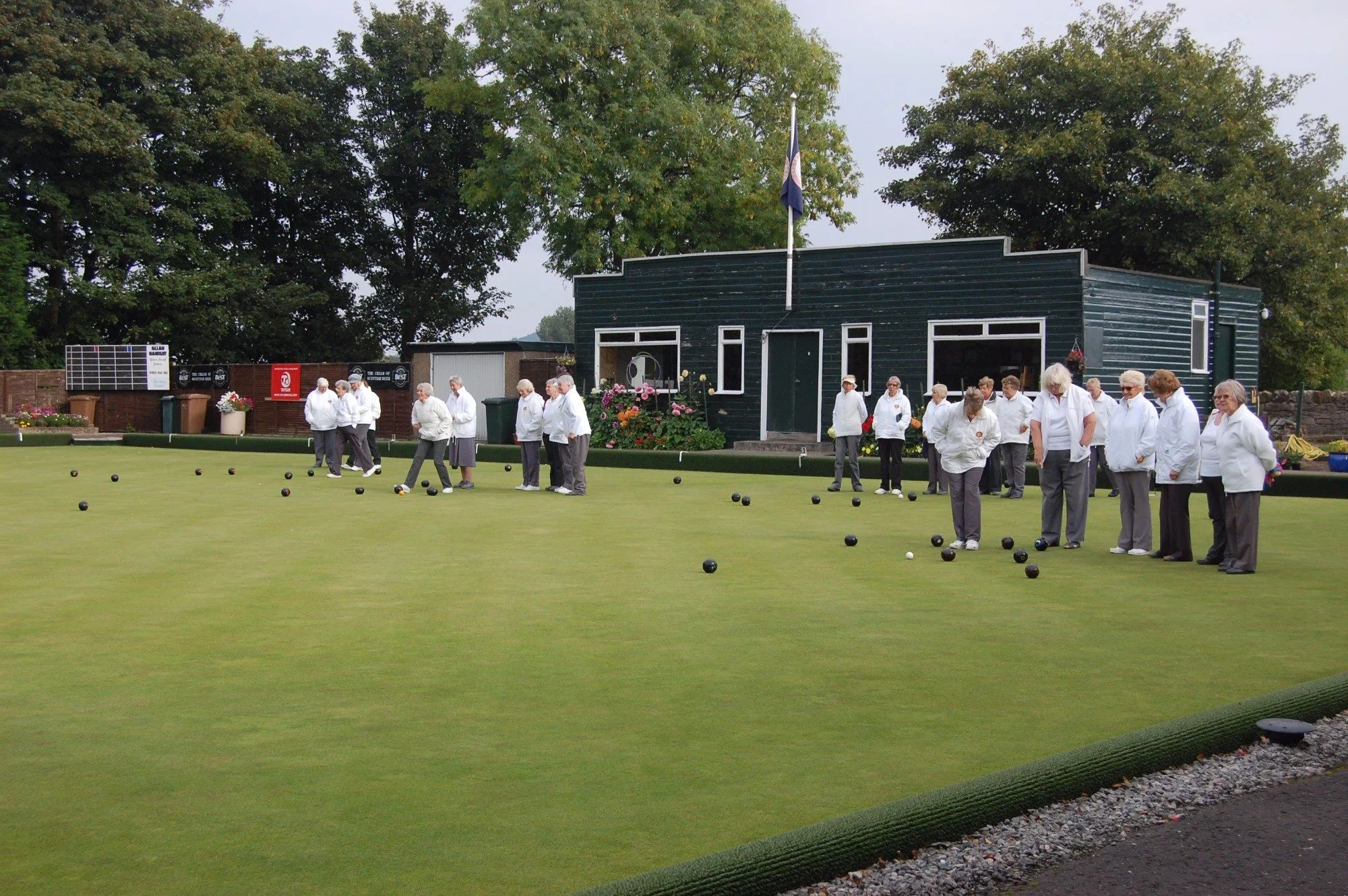 A group of elderly people dressed in white jackets playing or practicing bowls on a green outdoor lawn surrounded by trees.
