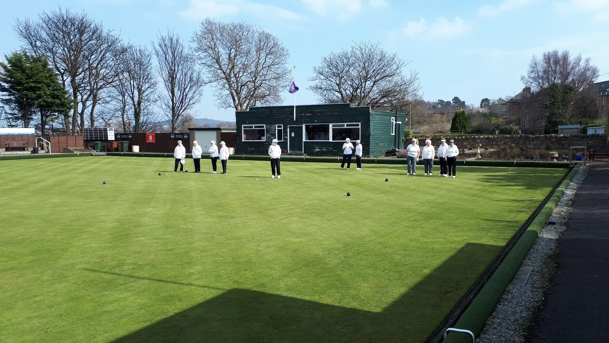 Group of older women playing lawn bowls on a green field outdoors, with a green building and trees in the background.