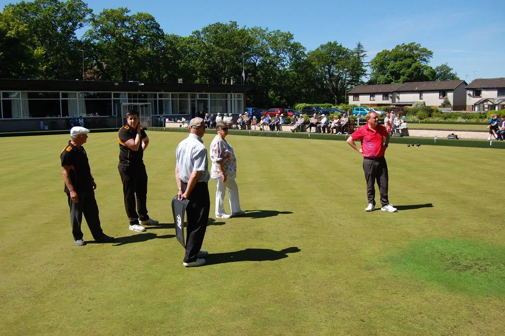 People preparing on a lawn bowling green with building and trees in the background.