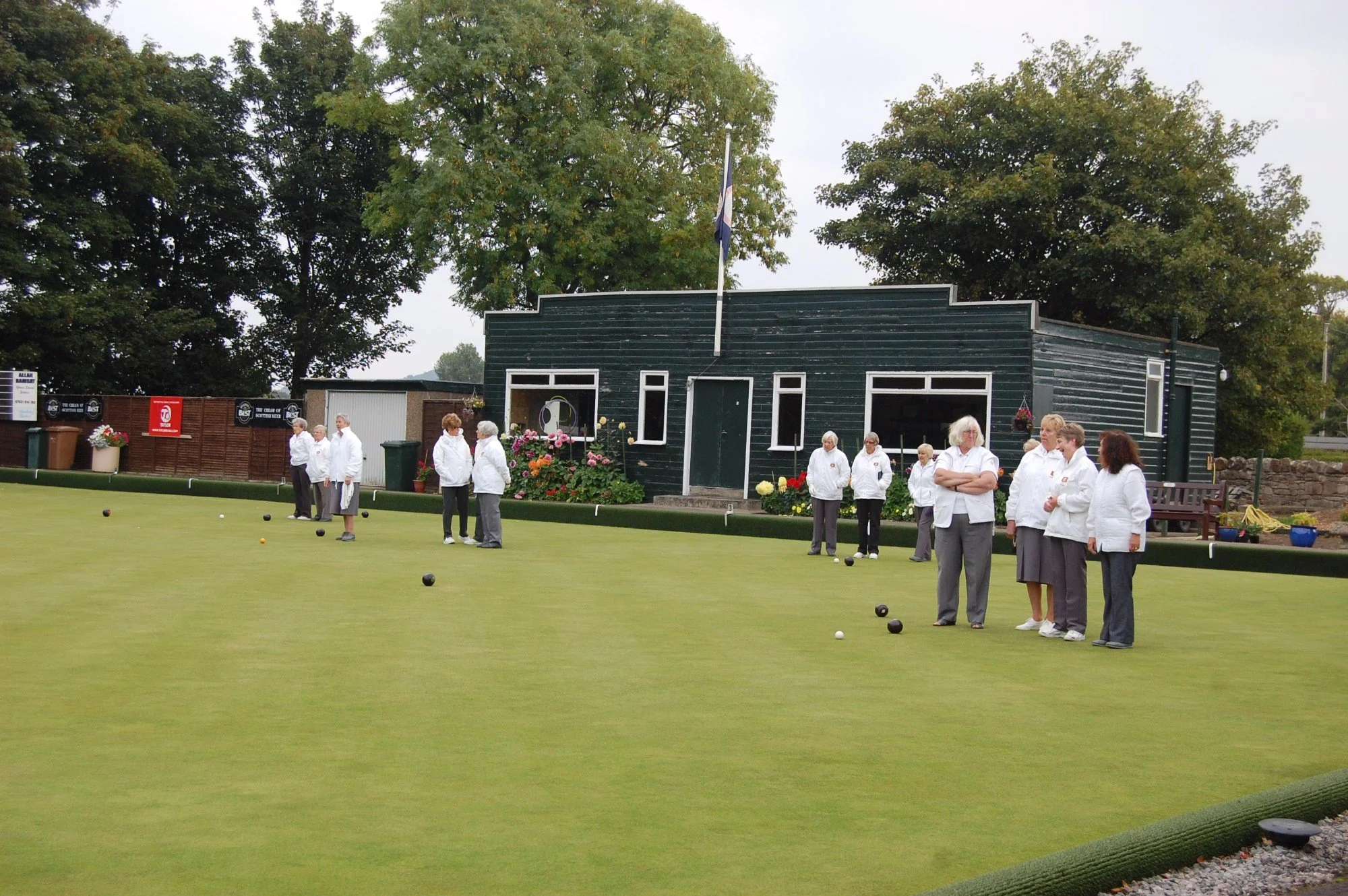 Group of older women playing or preparing to play lawn bowls outdoors, wearing white jackets and standing on a green playing field with trees and a building in the background.