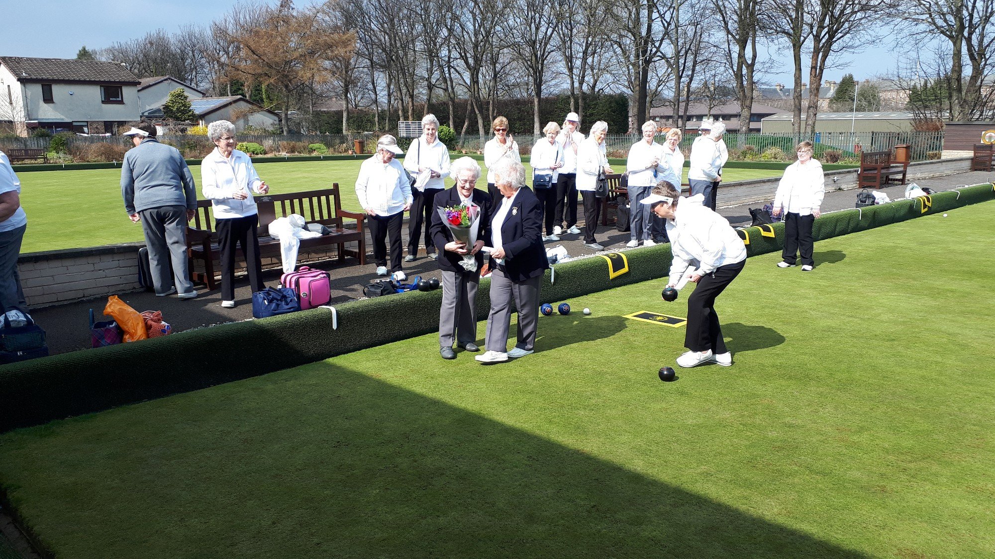 A group of elderly women playing lawn bowls outdoors on a sunny day, with some standing and others preparing to bowl.