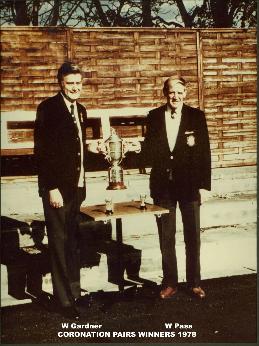 Two men standing outdoors in front of a wooden fence, each wearing dark suits with medals. They are holding a large trophy together, with a table between them that has a smaller trophy or cup. The photograph has a caption at the bottom that reads, "W