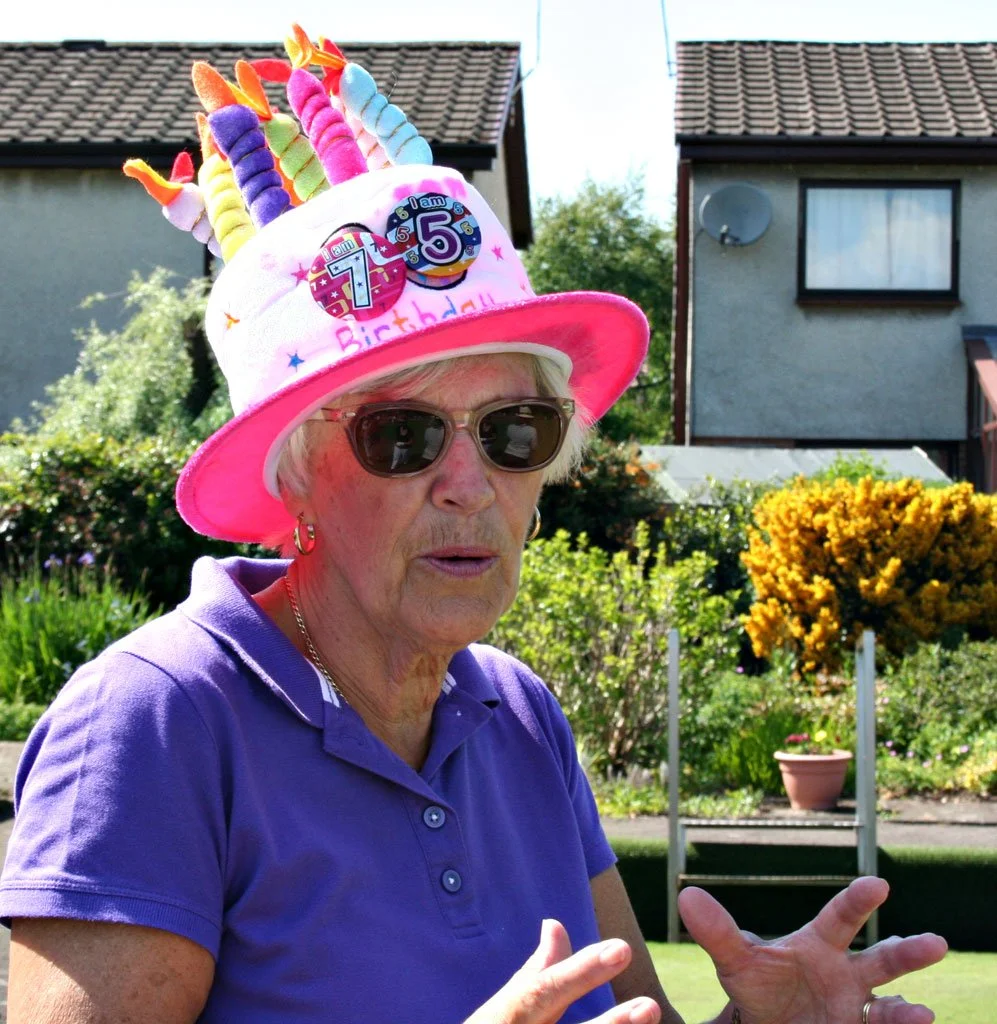 An elderly woman wearing sunglasses and a purple polo shirt outdoors, celebrating her birthday with a colorful birthday hat adorned with unicorn horns, candles, and the text 'I am 5' and '7'. She is gesturing with her hands in front of a garden and h