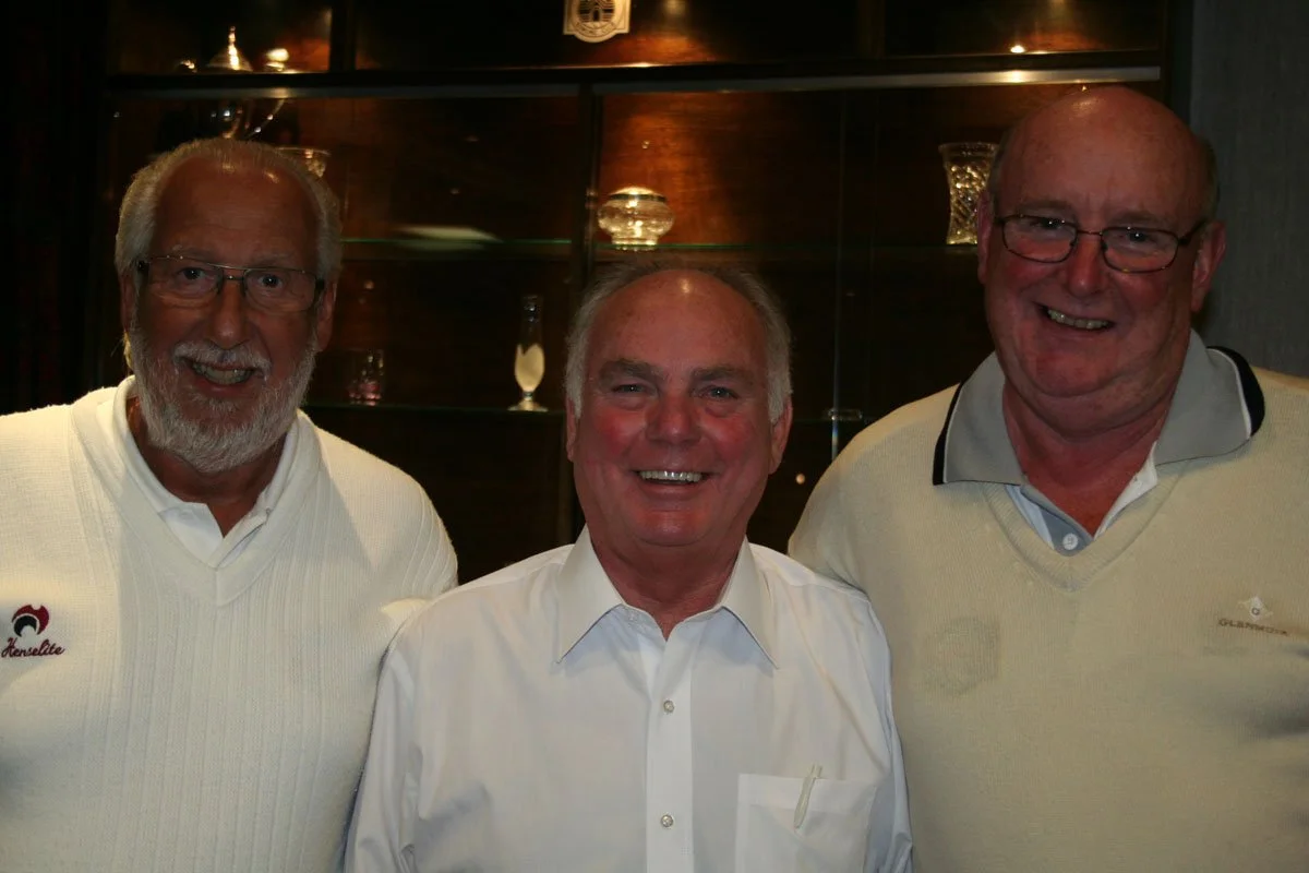 Three smiling older men standing close together indoors, behind a dark wooden display cabinet with glass shelves and vases.