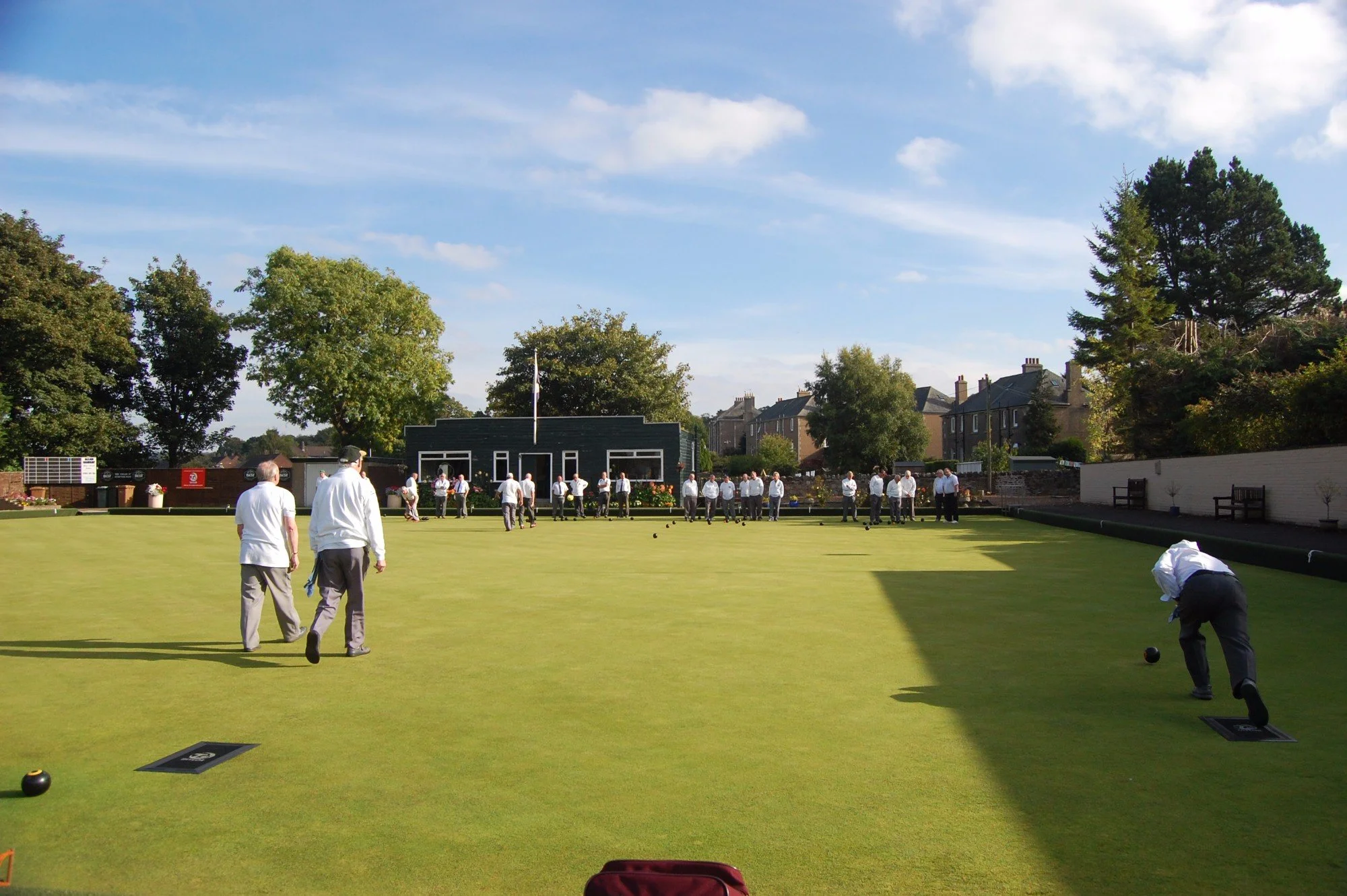A group of people playing lawn bowls on a neatly trimmed green on a sunny day, with trees and houses in the background.