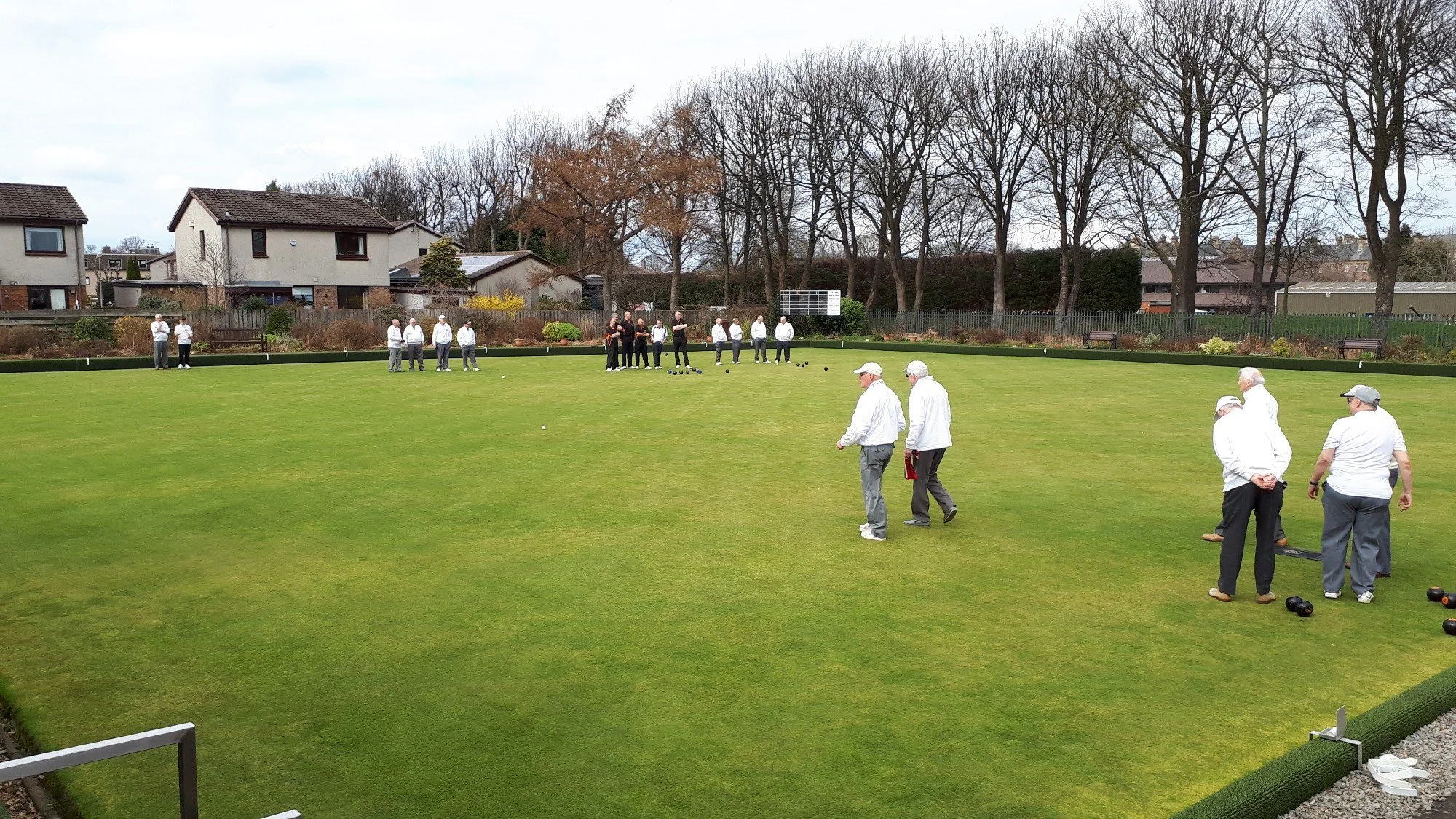 People playing lawn bowls on a green field surrounded by trees and residential houses.