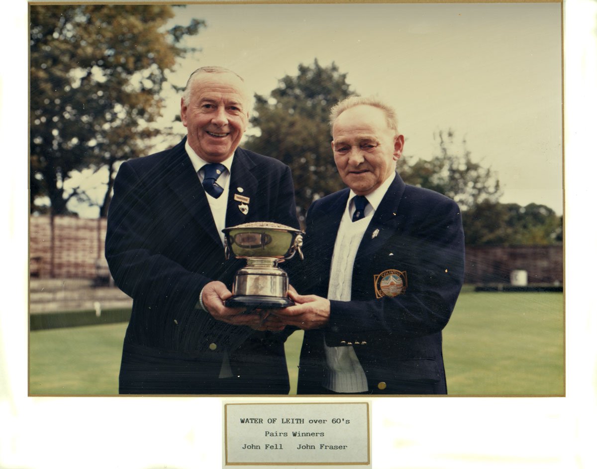 Two elderly men in formal suits and ties standing outdoors on grass, holding a trophy together, smiling. They are wearing badges and pins. The background shows trees and a fence, with an overcast sky. There is a plaque beneath the photo with the text