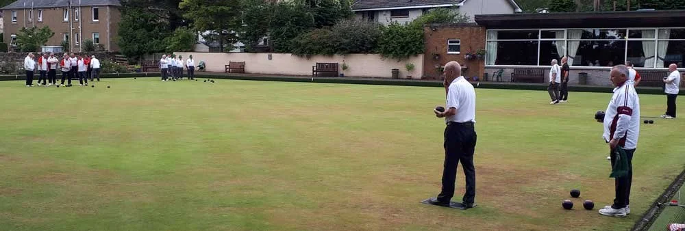 People playing lawn bowling on a green outdoor field, with some preparing to bowl and others observing, surrounded by buildings and trees.