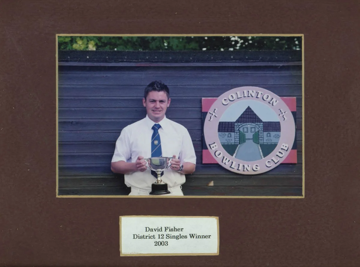 Young man in white shirt and tie holding a trophy, standing in front of a building with a sign that reads 'Collinton Bowling Club'