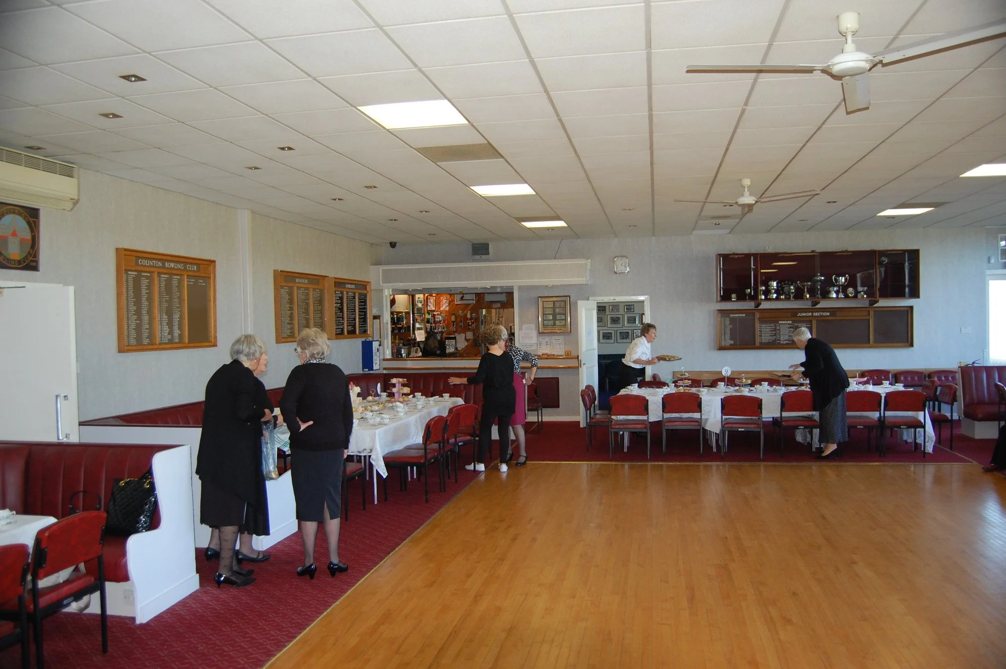 A group of mostly older women dressed in formal attire inside a bowling club's dining area, some standing and chatting while others prepare or serve food, with tables set with plates and cups, and a bar or service counter in the background.