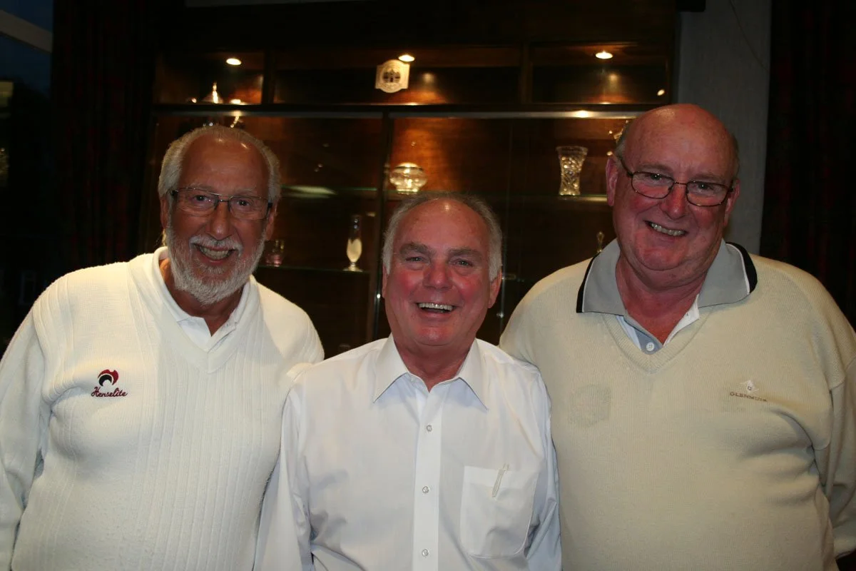 Three smiling men standing together indoors, with a glass display case with glassware behind them.