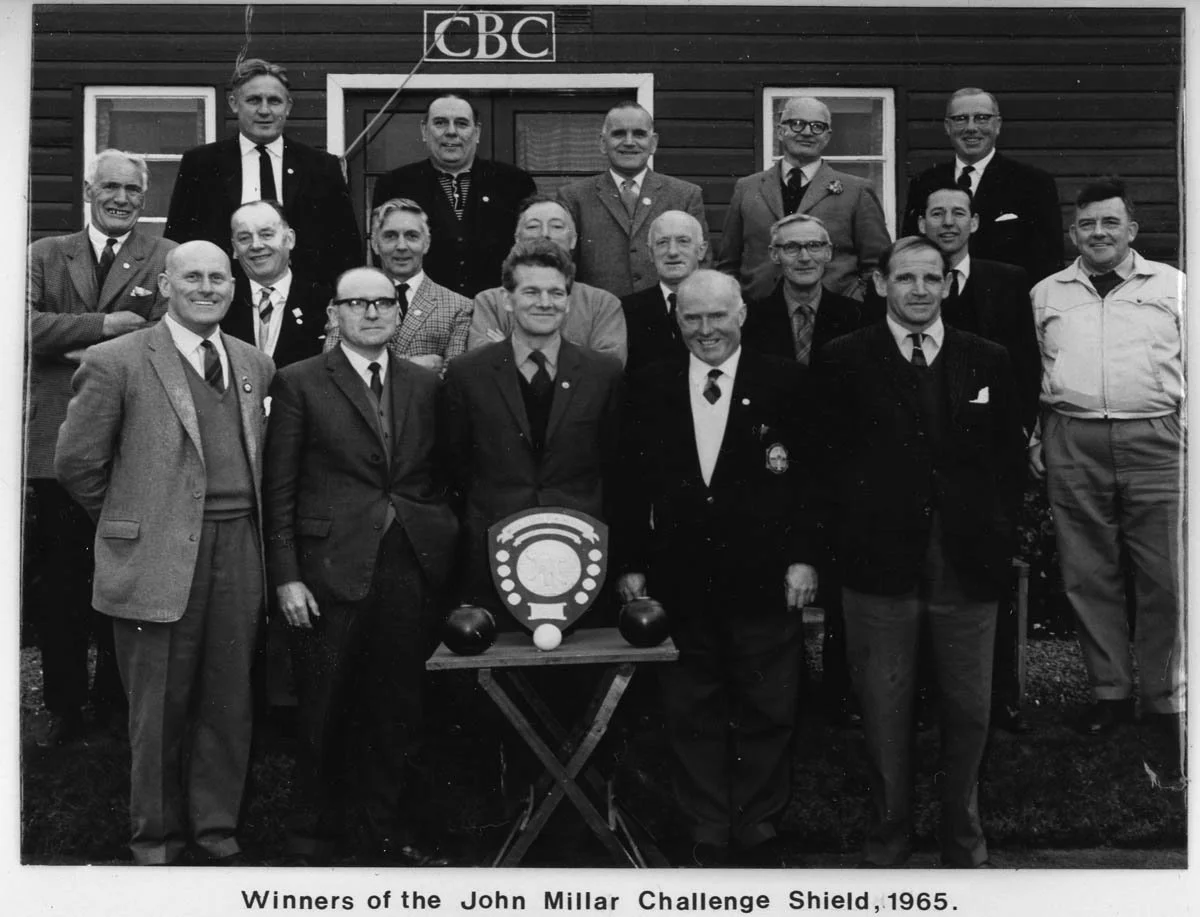 A black and white photo of 16 men standing outside in front of a wooden building with a sign labeled 'CBC'. They are dressed in suits, ties, and some in uniforms. There is a table in front of them with a shield-shaped trophy and two bowls. The captio