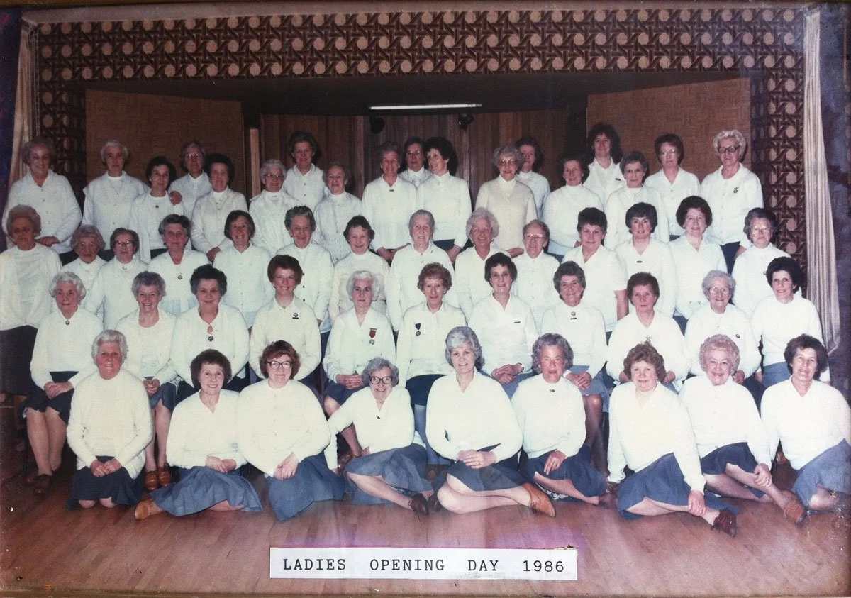 A group of women dressed in white blouses and dark skirts posing for a photo on Ladies Opening Day in 1986. They are arranged in rows on a stage with a wooden backdrop, in an indoor setting.