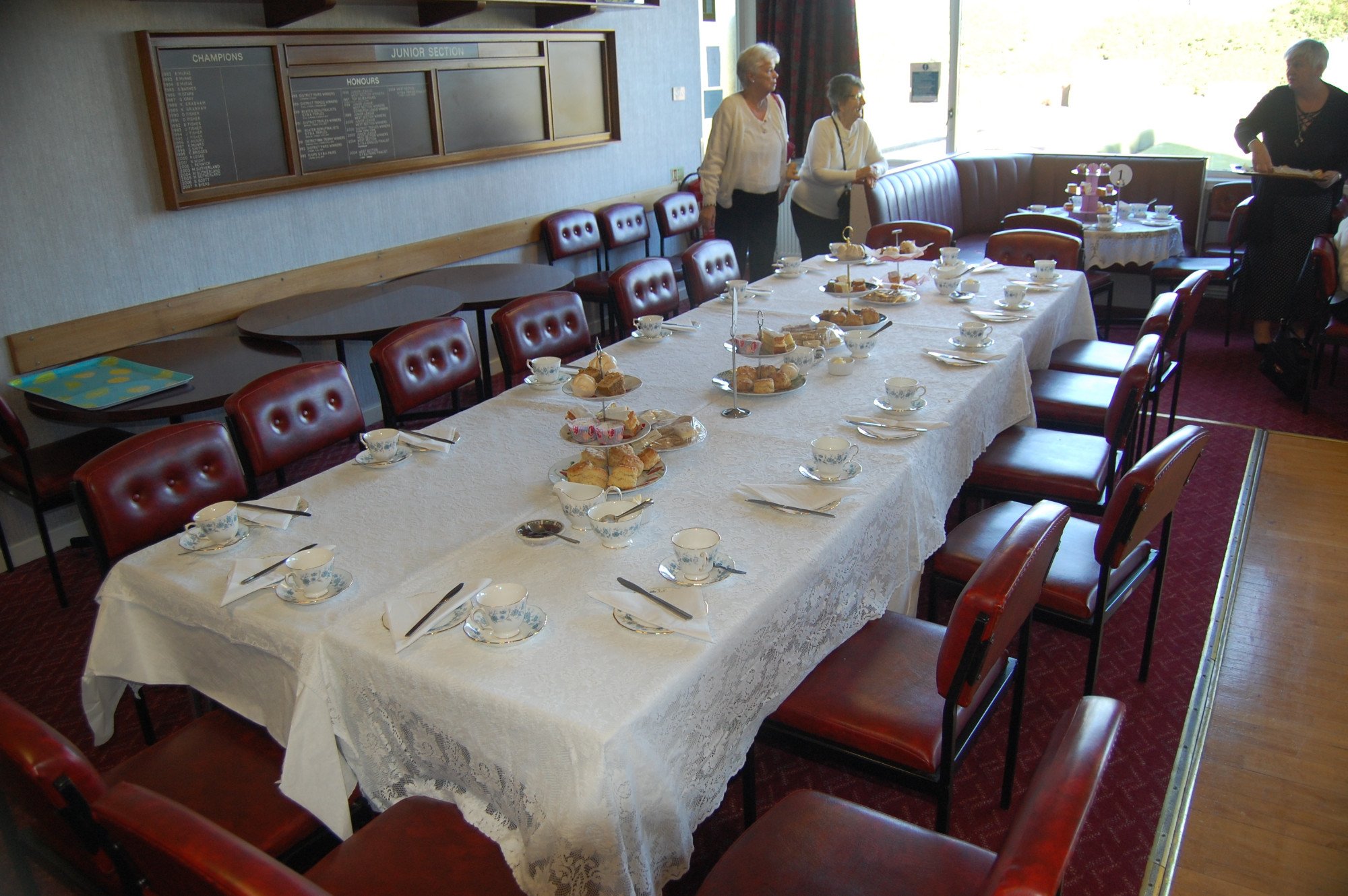 A dining room with a long table covered with a white lace tablecloth set for a tea party, featuring tea cups, plates with scones and sandwiches, and tiered trays with desserts. There are four women standing and conversing near the table, with additio
