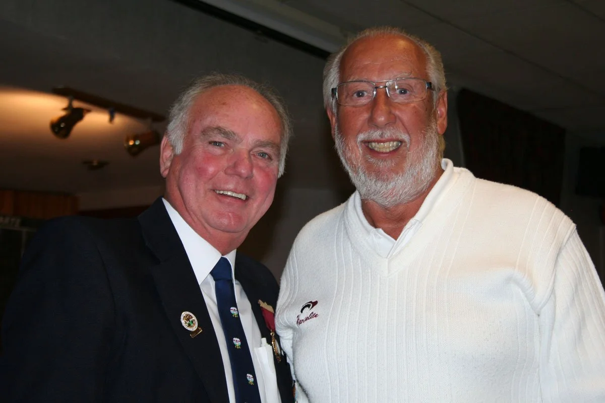 Two older men smiling and standing close together indoors. The man on the left is wearing a suit with pins and a tie, while the man on the right is wearing glasses and a white sweater.