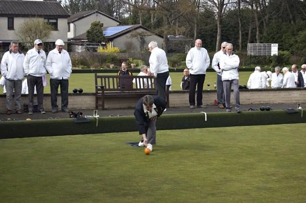 Person bending down to place a bocce ball on a bocce court surrounded by spectators, some wearing white jackets, with houses and trees in the background.