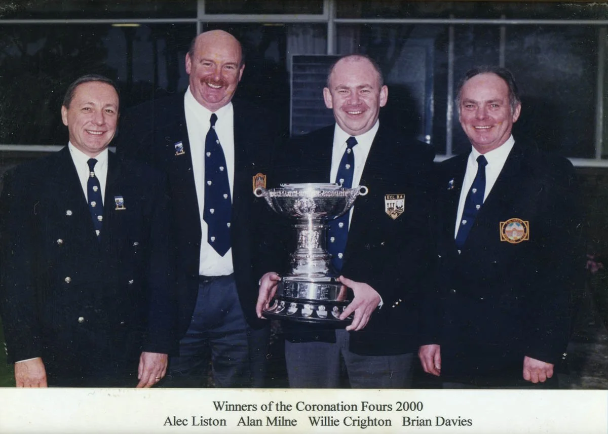 Five men in formal suits and ties standing together, holding a large trophy. They are smiling and appear to be celebrating, with a building in the background. The caption indicates they are the winners of the Coronation Fours 2000, with their names l