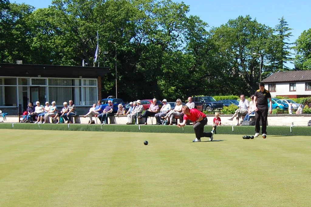 People playing lawn bowls on a green with spectators seated behind them and a building and parked cars in the background.