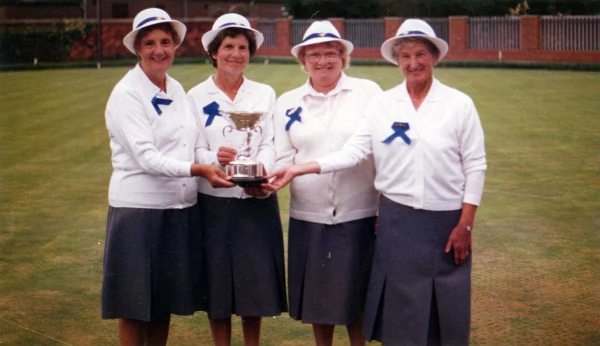 Four women in white shirts and dark skirts, wearing white hats with blue ribbons, standing on a lawn and holding a large trophy together, smiling.
